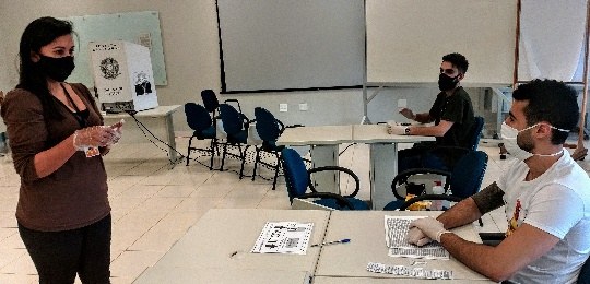 TRE-PR realiza simulação de votação com EPIs Fotografia de uma seção eleitoral. Há uma cabine de votação, mesários e eleitores com máscara
